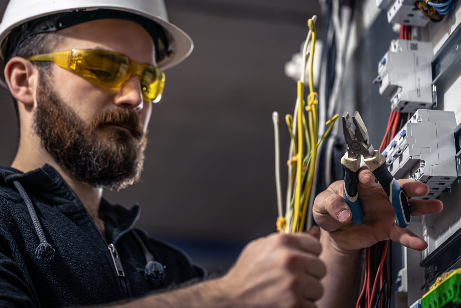 A male electrician works in a switchboard with an electrical connecting cable, connects the equipment with tools.